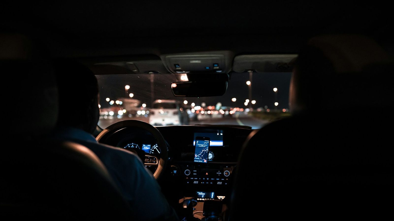 Zicht vanaf de achterbank van een auto bij nacht, de chauffeur als silhouet tegen de stadslichten